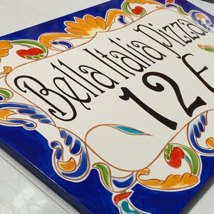 May include: A white ceramic sign with blue, green, yellow, and red floral designs. The sign says "Bella Italia Pizza" and "12E".
