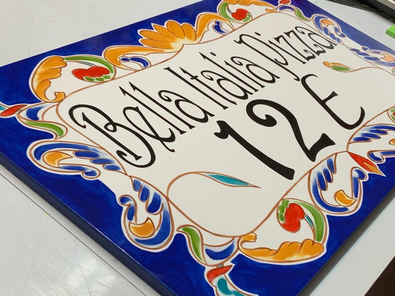 May include: A white ceramic sign with blue, green, yellow, and red floral designs. The sign says "Bella Italia Pizza" and "12E".