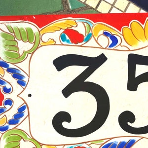May include: A rectangular ceramic house number plaque featuring the black digits 35 centered on a cream-colored background. The border is decorated with hand-painted floral and scroll motifs in vibrant shades of red, green, yellow, and blue.