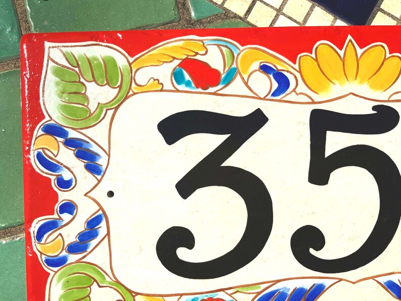 May include: A rectangular ceramic house number plaque featuring the black digits 35 centered on a cream-colored background. The border is decorated with hand-painted floral and scroll motifs in vibrant shades of red, green, yellow, and blue.