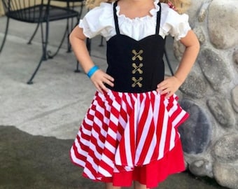 Pode incluir: Uma menina vestida de pirata para o Halloween. Ela está usando uma blusa branca, um colete preto com caveiras douradas, uma saia listrada vermelha e branca e botas pretas. Ela também está usando um tapa-olho e uma faixa de cabeça Minnie Mouse vermelha e preta com orelhas brilhantes.