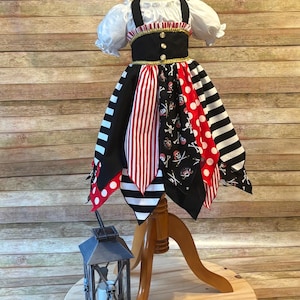 Op de afbeelding: Een piratenjurk voor kinderen op een houten mannequin. De jurk heeft een witte top met pofmouwen, een zwart lijfje met gouden accenten en een rok met rood-witte strepen, stippen en doodshoofdpatronen. Een decoratieve lantaarn staat in de buurt.