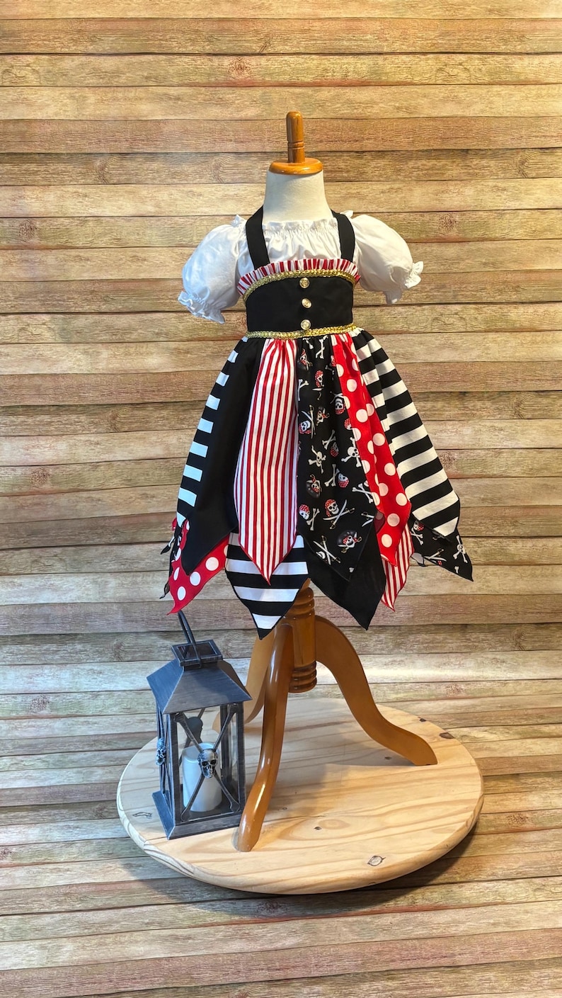 Op de afbeelding: Een piratenjurk voor kinderen op een houten mannequin. De jurk heeft een witte top met pofmouwen, een zwart lijfje met gouden accenten en een rok met rood-witte strepen, stippen en doodshoofdpatronen. Een decoratieve lantaarn staat in de buurt.