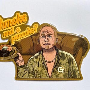 May include: A sticker featuring a cartoon man with a beard and a gold chain, sitting in a brown armchair. He is holding a plate with a pancake on it. The text "Shmoke and a pancake?" is written in a yellow, stylized font.