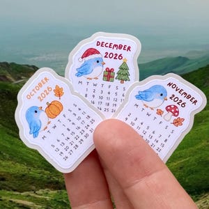 Puede incluir: Tres pegatinas troqueladas con las páginas del calendario de octubre, noviembre y diciembre de 2026. Cada pegatina muestra una ilustración de un pájaro azul, iconos estacionales como una calabaza, una seta y un árbol de Navidad.