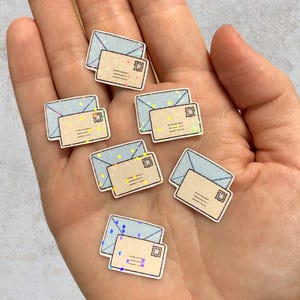 Op de afbeelding: Een verzameling van zes kleine, platte bedels in de vorm van een envelop. Elke bedel heeft een lichtblauwe envelop als basis met een kleinere, beige envelop erbovenop, afgewerkt met een subtiele holografische glitterafwerking.