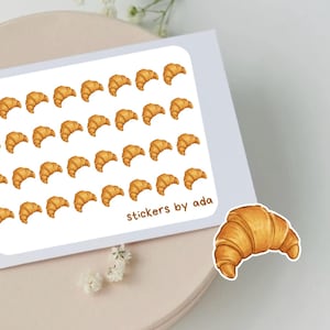 Op de afbeelding: Een rechthoekig stickervel met een herhalend patroon van kleine, goudbruine croissantillustraties. De tekst stickers by ada staat in de rechterbenedenhoek gedrukt. Een enkele, grotere croissantsticker is apart onderaan geplaatst.