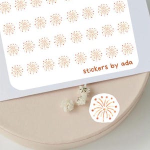 Può includere: Un foglio di adesivi rettangolare bianco con un motivo ripetitivo di piccole illustrazioni di fuochi d'artificio o stelle color rame disposte in file. Il testo stickers by ada è stampato in un piccolo carattere marrone in basso a destra.