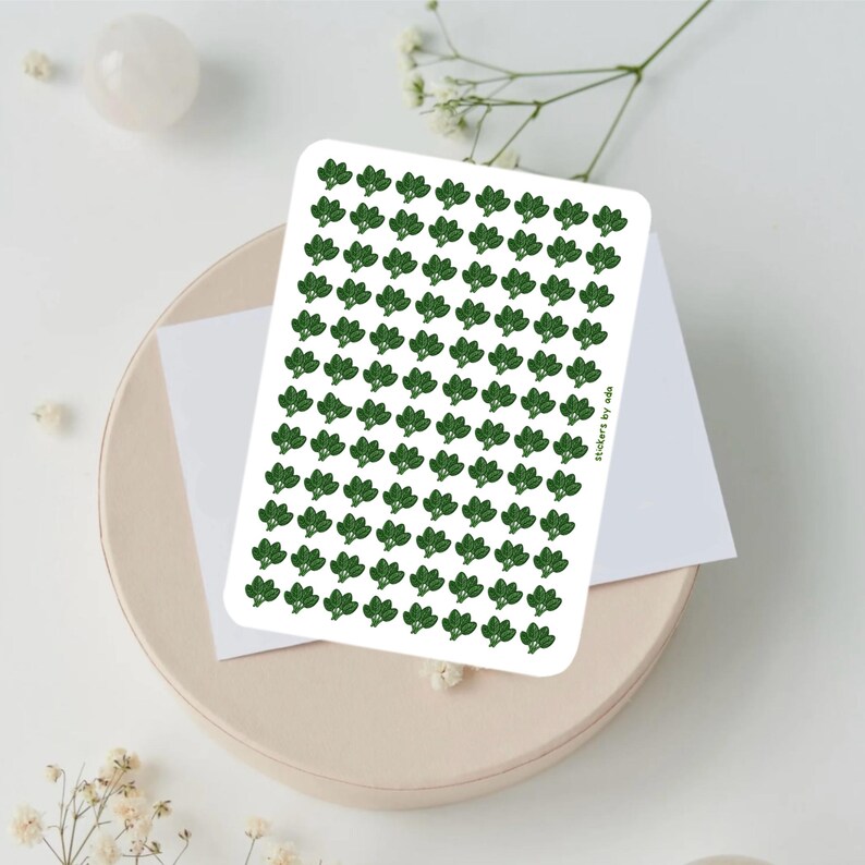 May include: A rectangular sticker sheet featuring a repeating pattern of small, dark green broccoli florets arranged in neat rows and columns. The text stickers by ada is printed in small, dark green letters along the right side of the sheet.