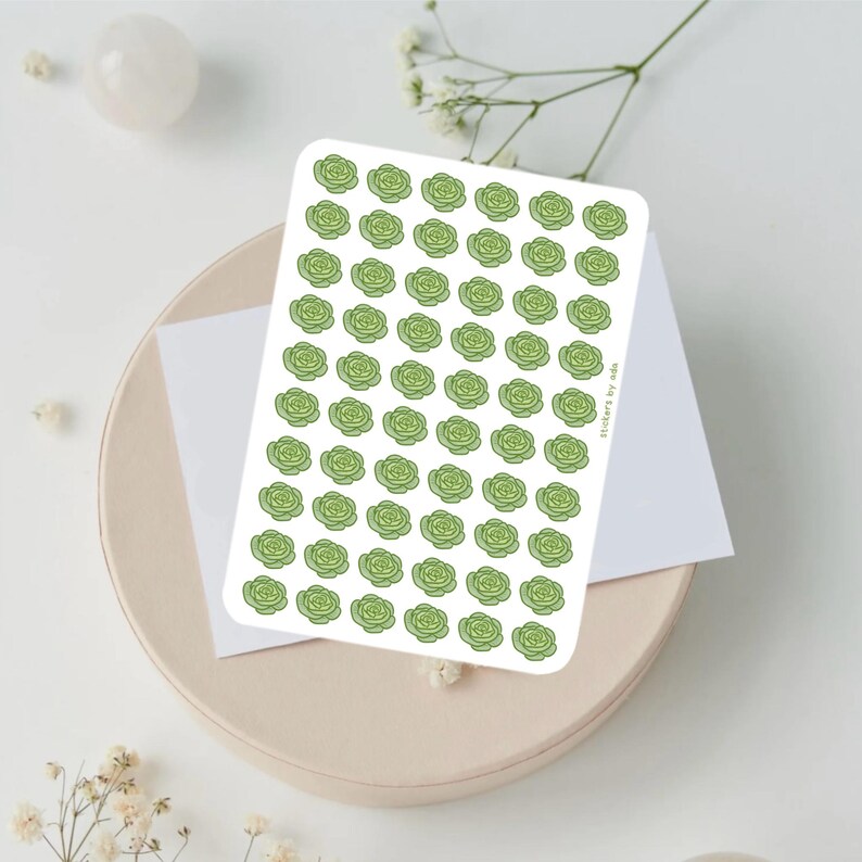 May include: A sheet of stickers featuring a pattern of light green rose illustrations. The stickers are on a white background, with the text "stickers by ada" visible. The sheet is placed on a beige plate with a white card.