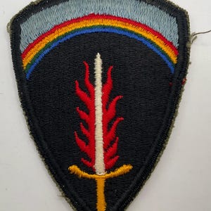 May include: A shield-shaped embroidered patch featuring a black background with a central vertical white sword blade surrounded by red flames and a yellow hilt. An arched rainbow band sits above the sword against a light blue sky-like backdrop.