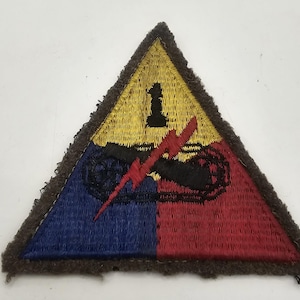 May include: A triangular embroidered patch featuring a yellow top section with a black number 1, a blue bottom left section, and a red bottom right section. A black tank tread graphic is overlaid with a red lightning bolt across the center.