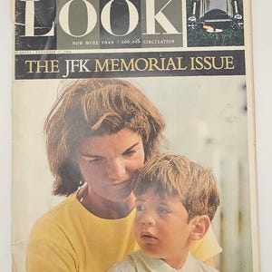 Pode incluir: Uma revista Look vintage de 17 de novembro de 1964, intitulada The JFK Memorial Issue. A capa apresenta uma fotografia de uma pessoa de cabelos castanhos vestindo uma camisa amarela segurando uma criança pequena com camisa branca de gola.
