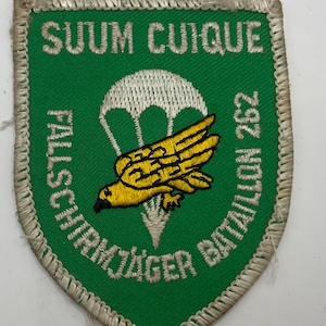 Puede incluir: Un parche bordado en forma de escudo con fondo verde y borde blanco. Presenta un paracaídas blanco sobre un pájaro amarillo, con el texto SUUM CUIQUE en la parte superior y FALLSCHIRMJÄGER BATAILLON 262 bordado en blanco a los lados.