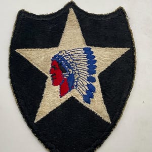 May include: A shield-shaped embroidered patch featuring a black background with a large, centered beige star. Inside the star is a profile illustration of a person wearing a traditional feathered headdress in shades of red, white, and blue.