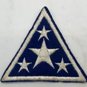 May include: A triangular embroidered patch featuring a dark blue felt background with a white stitched border. The design includes four white embroidered stars of varying sizes arranged in a triangular pattern across the center of the patch.