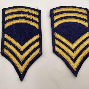 May include: A pair of matching military rank patches featuring a dark blue fabric base with gold embroidered stitching. Each patch displays three curved horizontal bars at the top and three downward-pointing chevrons at the bottom.