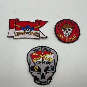 May include: Three embroidered patches. One is a red and white flag with a skull wearing a blue hat and crossed swords. One is a circular red patch with a skull in a conical hat and the text CAMBODIA VIETNAM LAOS SAT-CONG. One is a grey skull patch.