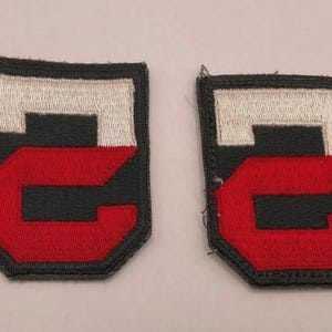 May include: Two rectangular embroidered patches featuring a black background with a white horizontal bar at the top and a large red letter C shape below. The patches have a dark border and a shield-like shape with a flat top and angled bottom.