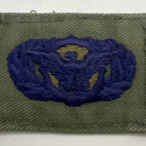 Puede incluir: Un parche rectangular de tela color verde oliva con un diseño bordado en hilo azul oscuro y dorado. El emblema central representa un águila estilizada con las alas extendidas, rodeada por un patrón de corona circular sobre la base.