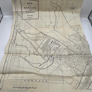 May include: A vintage paper map titled Map of Bangor and Vicinity Maine, featuring a grid layout with letters A through F and numbers 0 through 9. The map displays Dow Air Force Base, local roads, and the Penobscot River in black ink on cream paper.