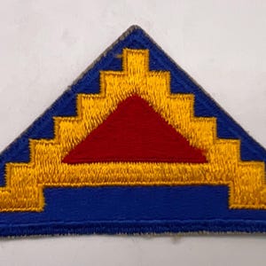 May include: A triangular embroidered patch featuring a blue background with a central red triangle surrounded by a yellow stepped border. The design consists of a solid blue field, a smaller red triangle in the center, and a yellow zigzag pattern.