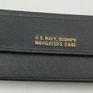 May include: A rectangular black textured case with a flap closure secured by two dark buttons. The front features gold lettering that reads U.S. NAVY, BUSHIPS NAVIGATOR'S CASE. The case has a structured, elongated design with a pebbled surface finish.