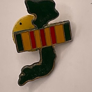 Könnte beinhalten: Eine Anstecknadel aus Metall mit einer stilisierten grünen Form, die an einen Vogel oder eine abstrakte Figur erinnert, akzentuiert durch einen horizontalen rechteckigen Balken. Der Balken zeigt abwechselnde vertikale Streifen in dunkelgrün, gelb und rot.
