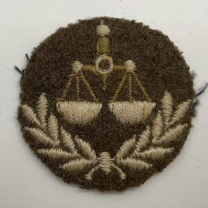 May include: A circular olive green felt patch featuring an embroidered design of a scale of justice centered above a pair of crossed laurel leaves. The embroidery is executed in shades of light tan and beige thread, creating a textured appearance.