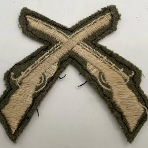 May include: An embroidered fabric patch featuring two crossed flintlock pistols in tan thread against a dark olive green felt background. The design shows detailed stitching for the barrels, triggers, and grips of the firearms in a cross formation.