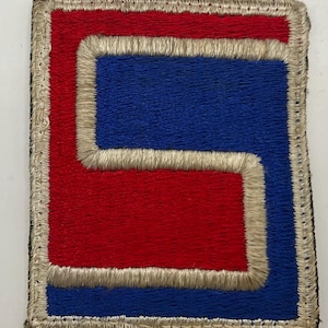 May include: A rectangular embroidered patch featuring a stylised S-shaped design. The patch is bordered in off-white thread, with a central red S-shape set against a blue background. The embroidery is dense, creating a textured surface with clear colour contrast between the red, blue, and off-white sections.