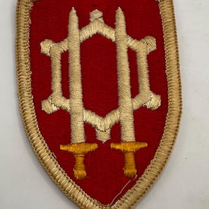 May include: A shield-shaped embroidered patch featuring a red background with a gold border. The center displays a gold-colored gear-like emblem intertwined with two vertical swords, with the sword hilts rendered in a distinct yellow thread.