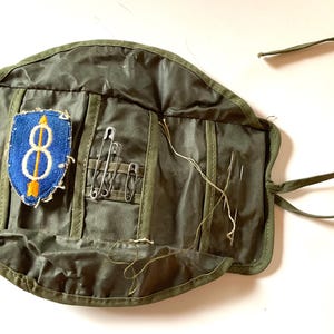 May include: An open, olive green canvas sewing kit with a blue and yellow patch, safety pins, needles, and thread. The kit is rolled out, revealing pockets for the sewing supplies. A green tie secures the kit.
