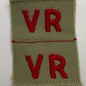May include: A rectangular olive green fabric patch featuring a diagonal twill weave. The patch is divided horizontally by a single red stitched line. The letters VR are embroidered in red thread in both the top and bottom sections of the patch.
