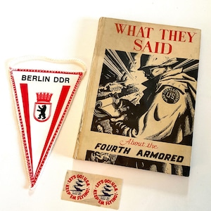 Puede incluir: Un conjunto de objetos vintage: un banderín con "BERLIN DDR" y un emblema de oso, un libro titulado "WHAT THEY SAID" con una ilustración de guerra, y dos pequeñas pegatinas "LET'S GO! USA" con gráficos de aviones.