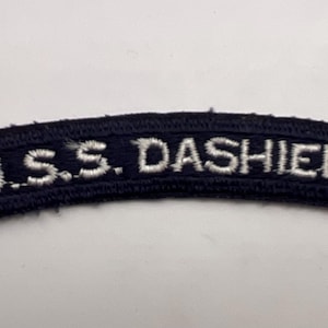 May include: A curved, dark navy blue embroidered patch featuring the white text U.S.S. DASHIELL stitched in a clean, sans-serif font. The patch has a finished border and a slightly arched shape, designed for attachment to a uniform or garment.
