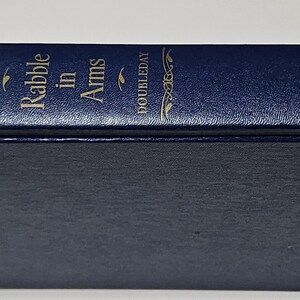 May include: A navy blue hardcover book titled "Rabble in Arms" by Kenneth Roberts. The book's spine features gold lettering and the publisher's name, Doubleday. The book has a textured cover.