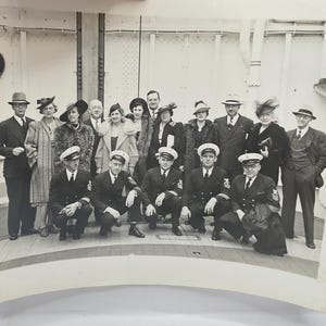 May include: A black and white vintage photograph showing a group of fifteen people posing on a ship deck. The group includes men in naval uniforms with peaked caps and civilians in formal suits, coats, and hats, standing and kneeling in rows.