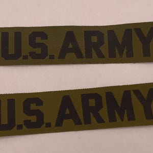 Könnte beinhalten: Zwei rechteckige olivgrüne Stoffaufnäher mit dem Text U.S. ARMY, der in fetten schwarzen Großbuchstaben aufgestickt ist. Die Aufnäher haben eine gewebte Textur und gerade Kanten und sind zur Befestigung an Kleidung oder Ausrüstung gedacht.