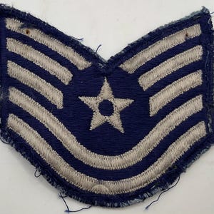 May include: A dark blue embroidered military chevron patch featuring four silver-colored stripes arranged in a V-shape with a central five-pointed star. The patch has a stitched border and shows signs of wear along the edges with loose threads.