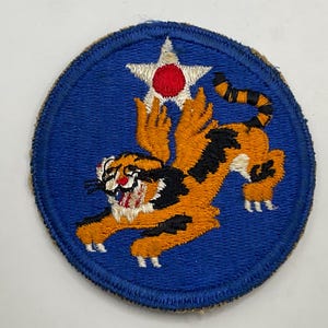 May include: A circular embroidered patch featuring a blue background with a leaping orange tiger with black stripes. Above the tiger is a white five-pointed star with a red circle in the center. The patch has a dark blue stitched border.