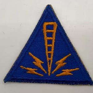 Könnte beinhalten: Ein dreieckiger gestickter Aufnäher mit einem leuchtend blauen Hintergrund, einem zentralen goldgelben vertikalen Turmsymbol und vier stilisierten Blitzsymbolen, die von der Basis des Turms ausgehen, abgerundet mit einem genähten Rand.