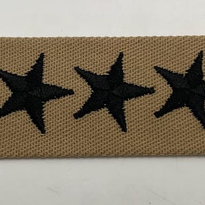 May include: A rectangular tan fabric patch featuring three embroidered black stars arranged horizontally in the center. The patch has a textured weave and is finished with thin black stitched borders on the far left and right edges.