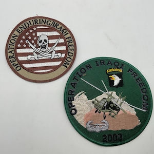 May include: Two circular embroidered patches. The top patch features a skull and crossbones over a red and white striped flag with the text Operation Enduring/Iraqi Freedom. The bottom patch is green with a helicopter, eagle, and 2003 text.