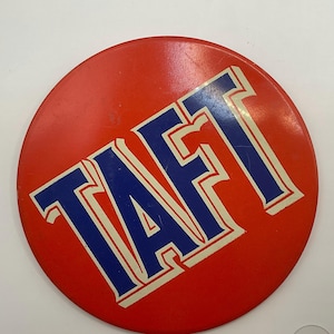May include: A circular red pinback button featuring the word TAFT in bold blue capital letters with a white outline. The text is positioned diagonally across the center of the button, which has a smooth, glossy finish and a metallic edge.
