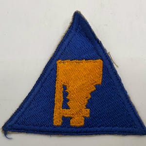 May include: A triangular embroidered patch featuring a vibrant blue background with a stitched border. Centered on the patch is a stylized, blocky letter A in a bright golden-yellow thread, creating a sharp contrast against the blue fabric.