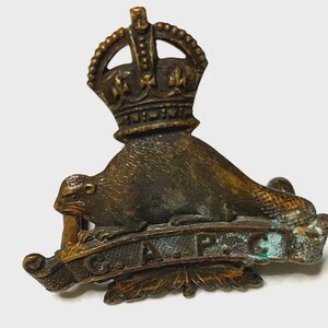 Puede incluir: Una insignia militar de metal en tono bronce que presenta un castor colocado sobre una cinta con las letras C.A.P.C. en relieve. El diseño está rematado con una corona e incluye detalles en forma de hojas en la base de la insignia.