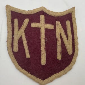 May include: A shield-shaped felt patch featuring a deep maroon background with a thick, beige border. The letters K, T, and N are appliqued in beige felt across the centre, with a cross symbol positioned between the letters K and T and N.