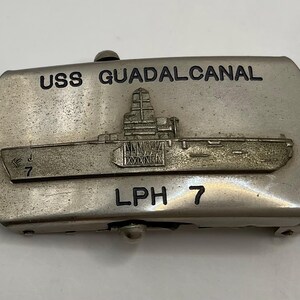 Pode incluir: Uma fivela de cinto retangular de metal prateado com um relevo de um navio de guerra. O texto USS GUADALCANAL está gravado no topo e LPH 7 na parte inferior. O número 7 também é visível na proa do navio.