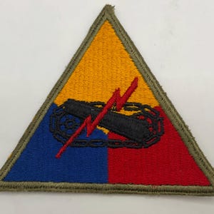 May include: A triangular embroidered patch featuring a yellow top section, a blue bottom-left section, and a red bottom-right section. Centered is a black tank tread graphic overlaid with a jagged red lightning bolt, all bordered in olive green.
